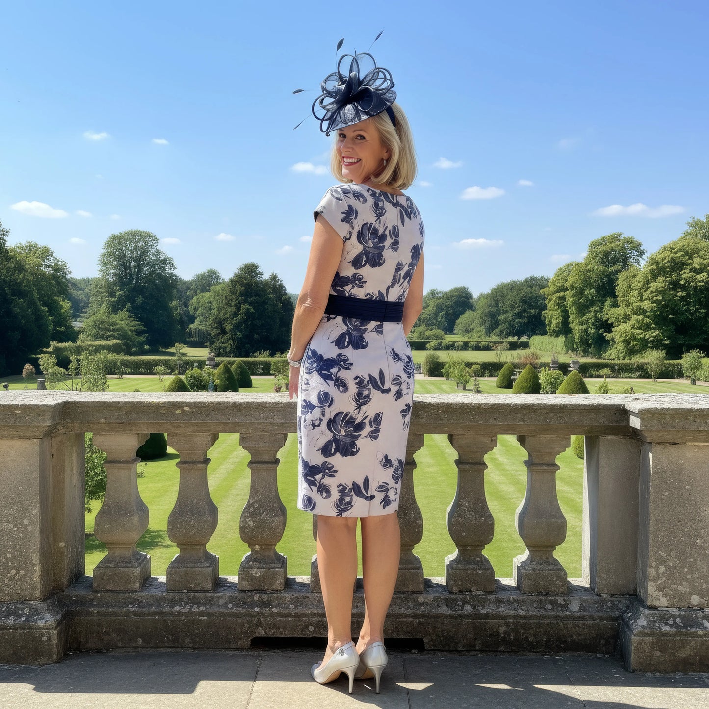 Woman in a floral dress with a hat standing on a stone balcony overlooking a garden.