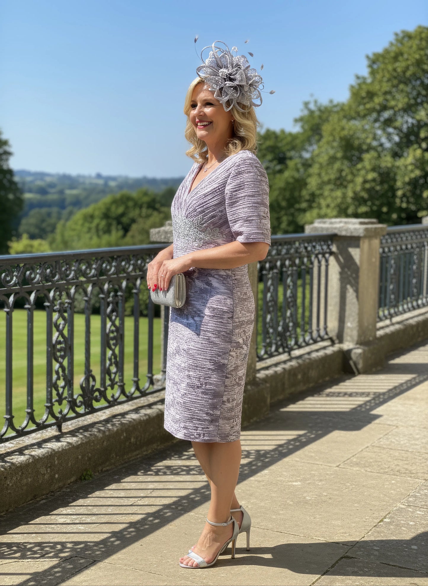 Woman in a patterned Condici grey dress as mother of the bride standing in front of a large building with greenery.