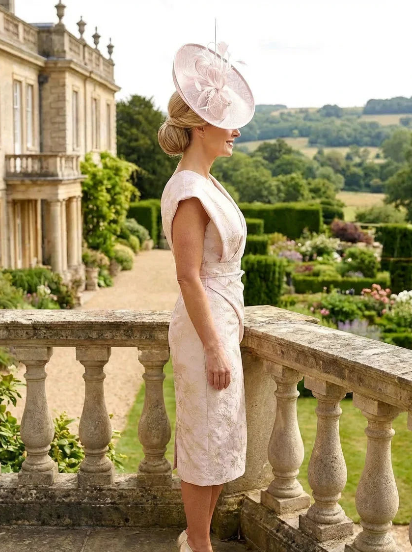 side view of a woman wearing a Gabriella Sanchez blush pink mother of the bride dress and matching blush pink fascinator stood on a balcony.