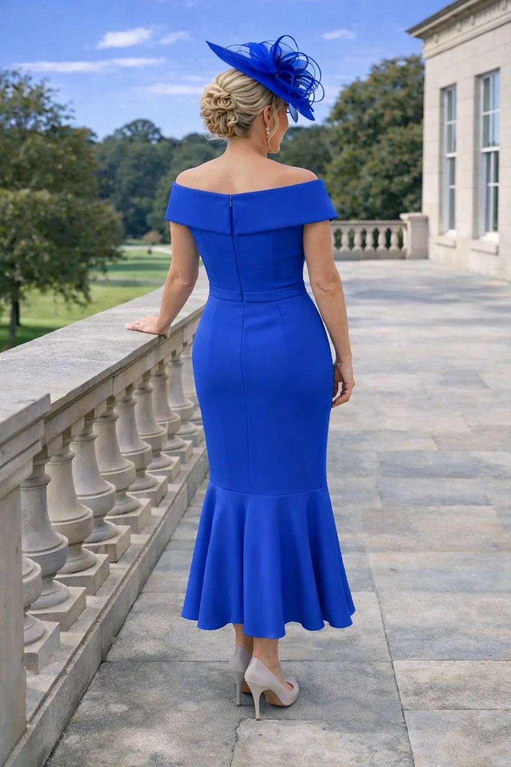 Back view of a woman in a Ispirato bardot cobalt blue mother of the bride dress and cobalt fascinator hat standing on a stone patio.