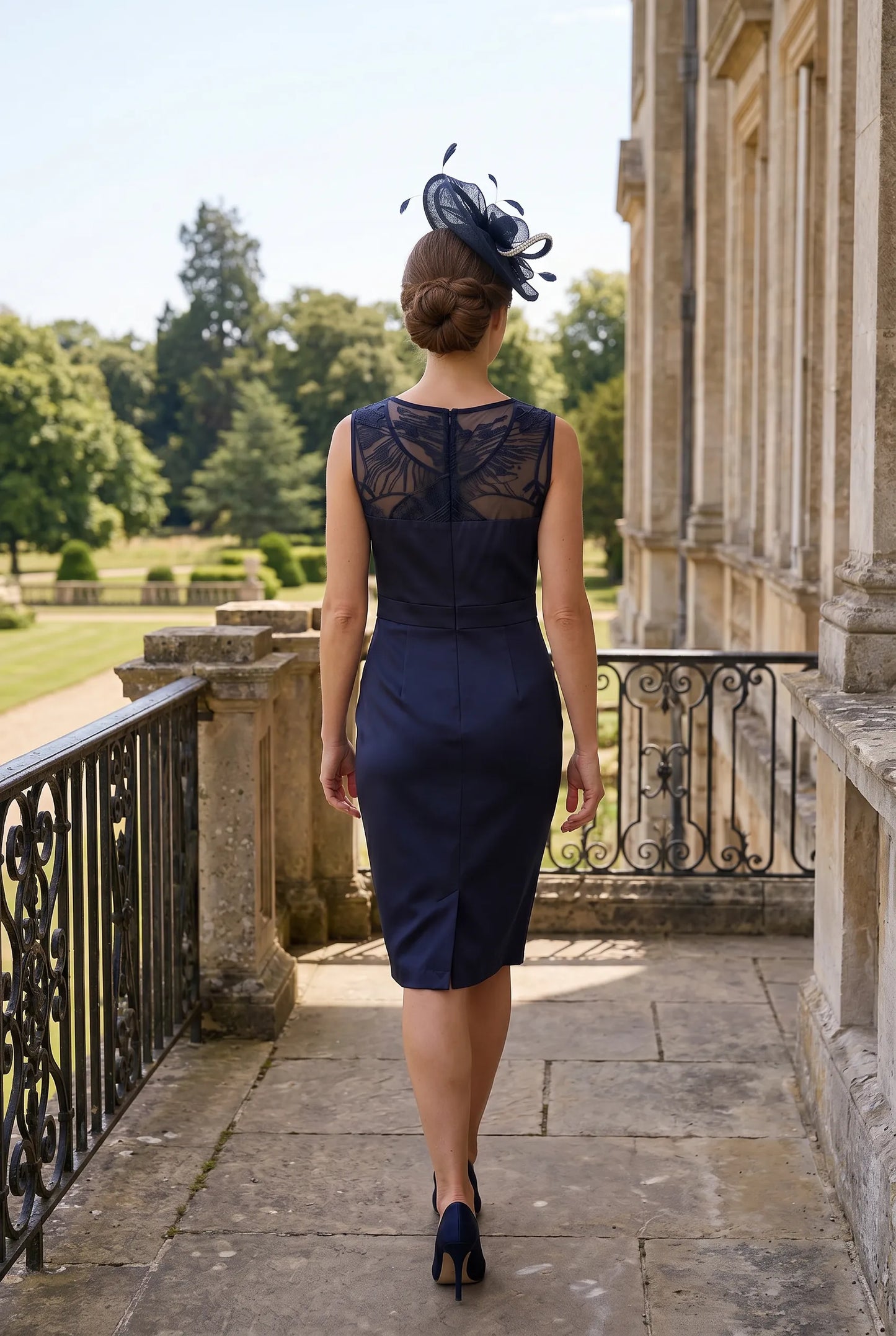 Woman in a navy dress with floral pattern and matching navy embellished oval fascinator walking on a stone path in an elegant outdoor setting.