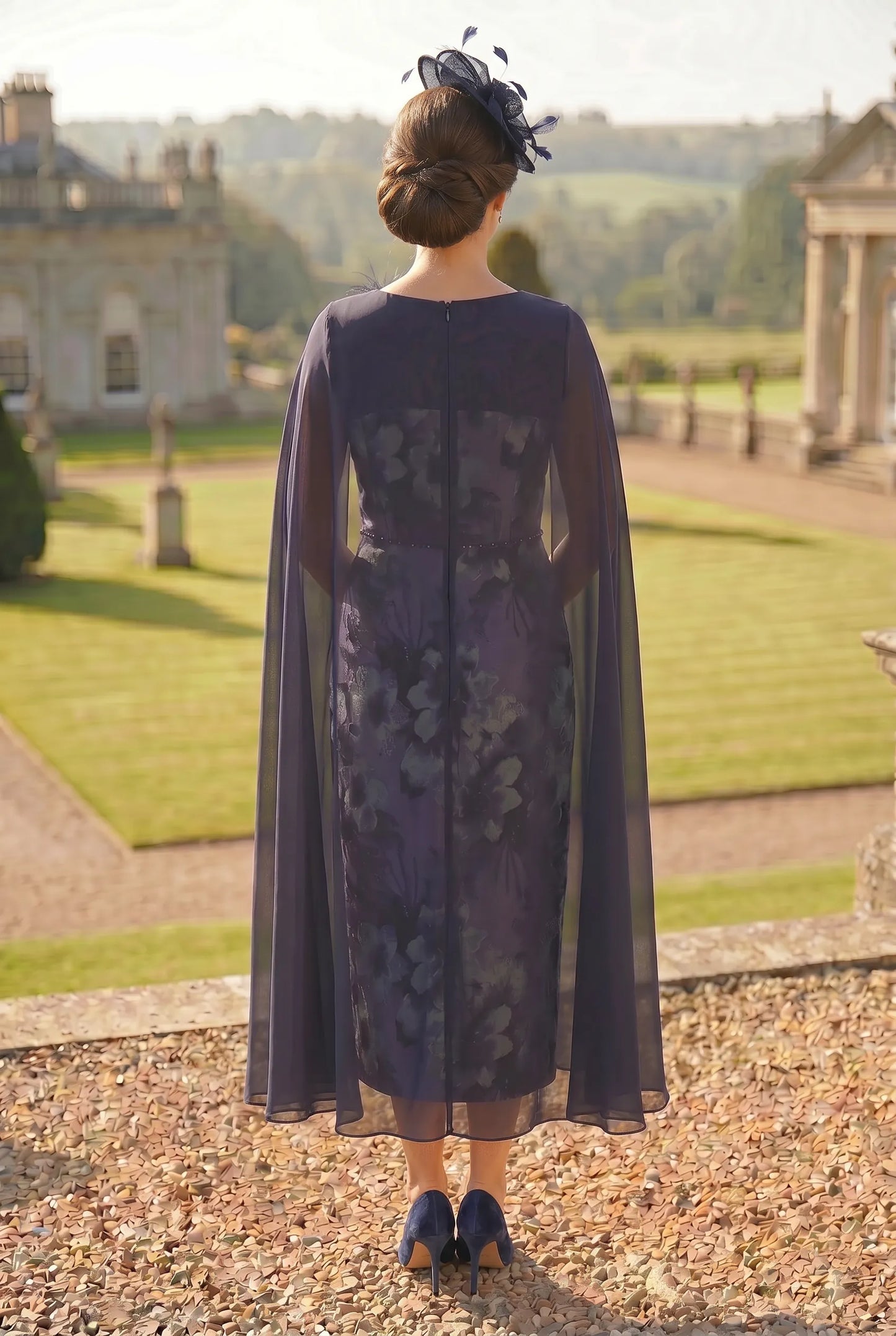 Woman in a dark floral dress with a long cape standing in a formal garden setting.