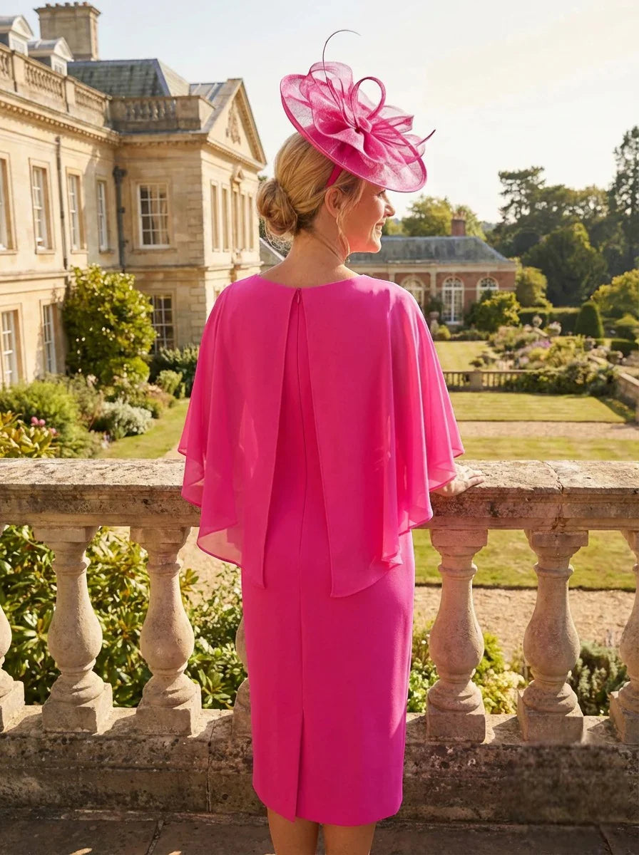 Back view of a woman in a Lizabella bright pink mother of the bride dress with chiffon cape sleeves and matching fascinator hat standing on a stone balcony with a garden view.