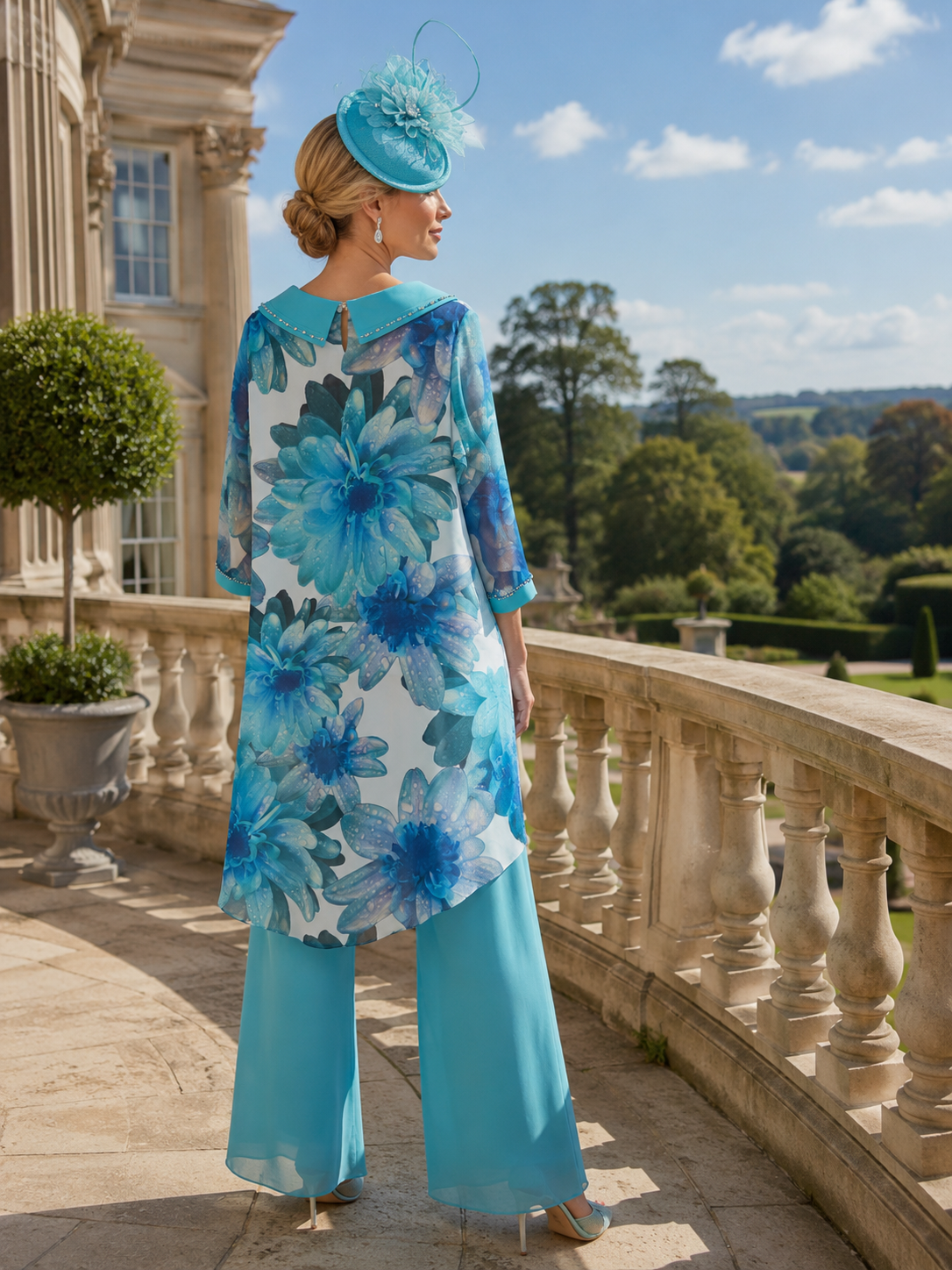 Woman in ablue floral mother of the bride 2 piece outfit and wearing a blue embellished fascinator wedding hat standing on a stone balcony with a scenic background.