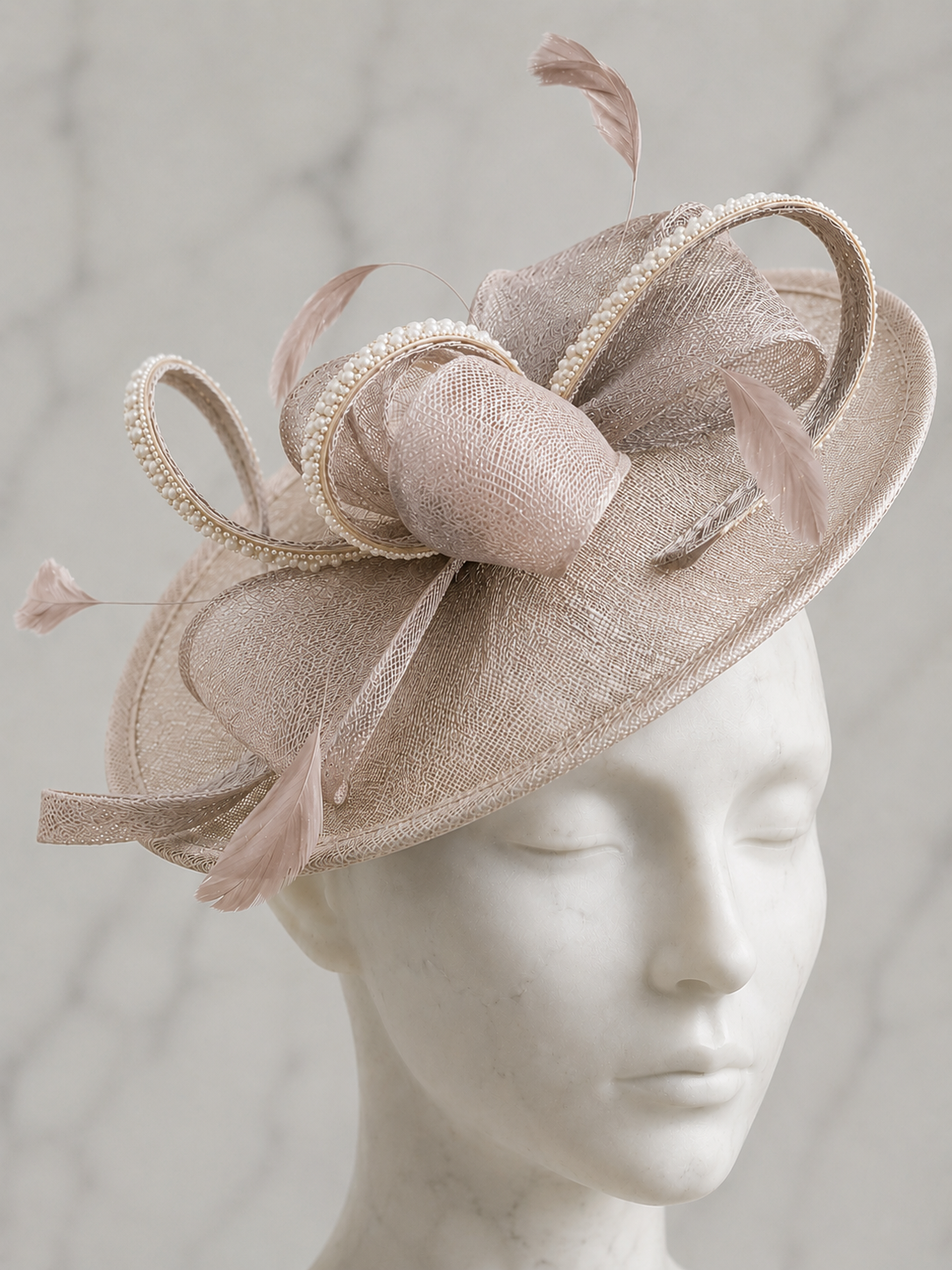 A Mother of the Bride pale taupe fascinator embellished hat with feathers, rhinestones and pearls sat on a white mannequin head.
