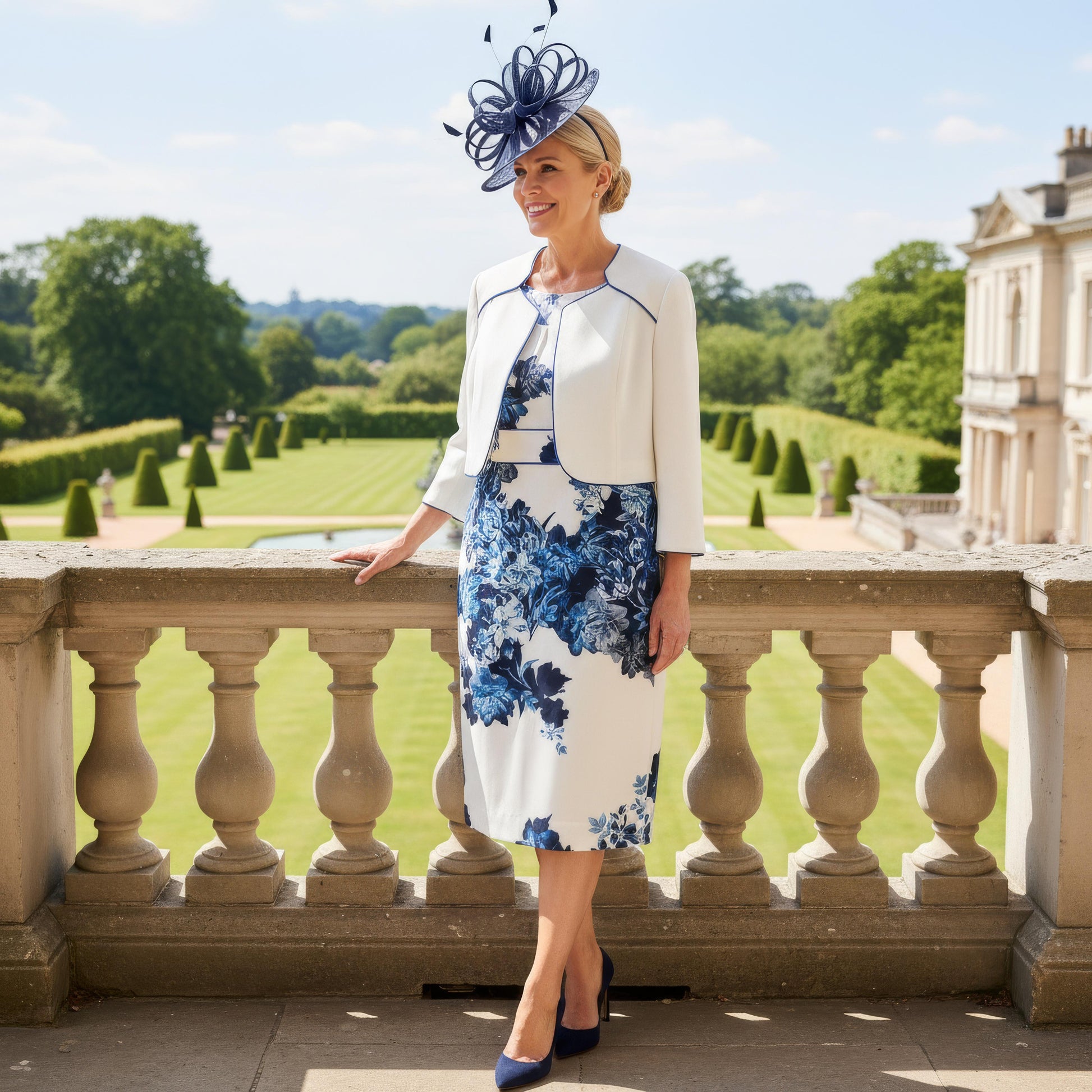 Elegant navy Mother of the Bride outfit by Condici 70820 featuring a fitted ruched dress with a coordinating cropped jacket, worn on a stone balcony overlooking manicured gardens.