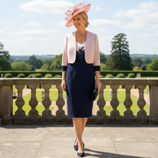 Woman in a Condici navy and pink dress and jacket and a pink fascinator hat standing on a stone patio with a scenic background. Signature Dress.