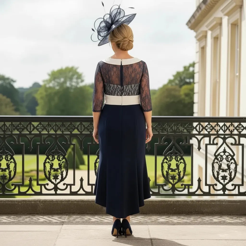 Back view of navy Couture Club dress with lace bodice, 3/4 sleeves and high‑low hemline, worn with a navy fascinator on a balcony.