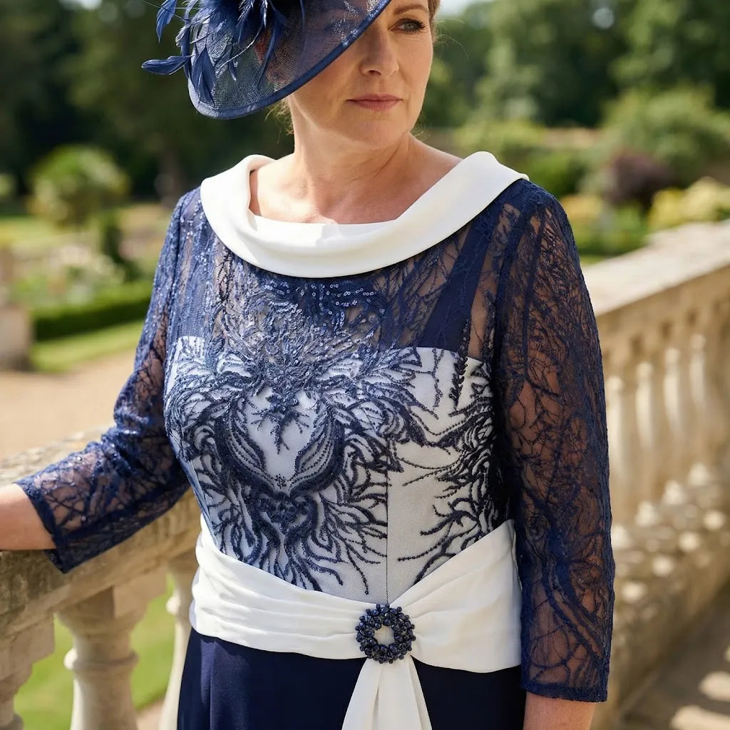 Front view of navy Couture Club high‑low dress with lace sleeves, cream waistband and brooch detail, styled outdoors with a navy fascinator.