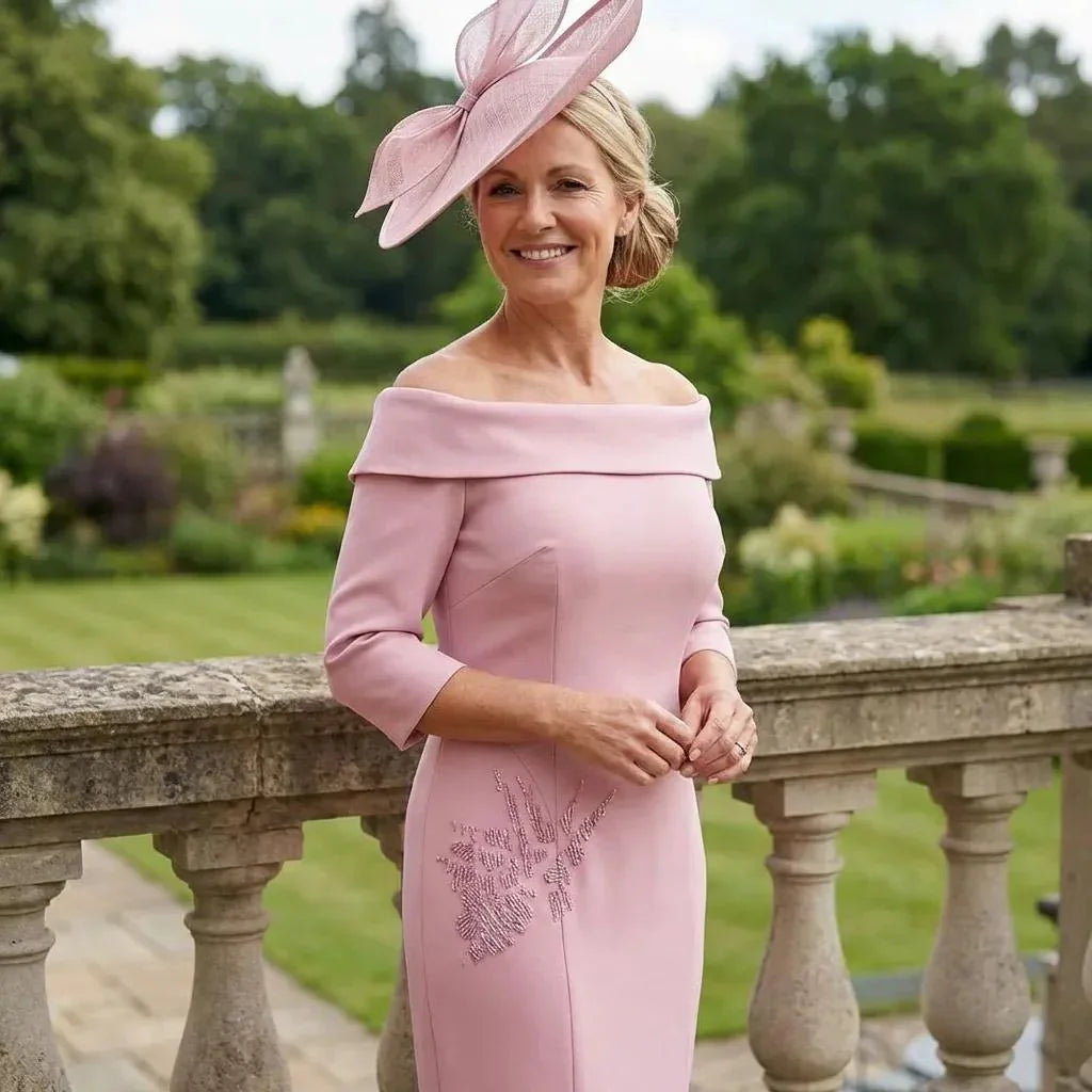 Front view of the Couture Club 8G150 blush pink off‑shoulder dress with subtle side embellishments and matching fascinator, photographed in a garden setting.