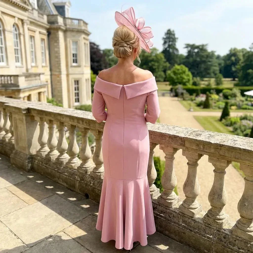 Back view of the Couture Club 8G150 blush pink dress with flared hem and matching fascinator, photographed on a terrace overlooking manicured gardens.
