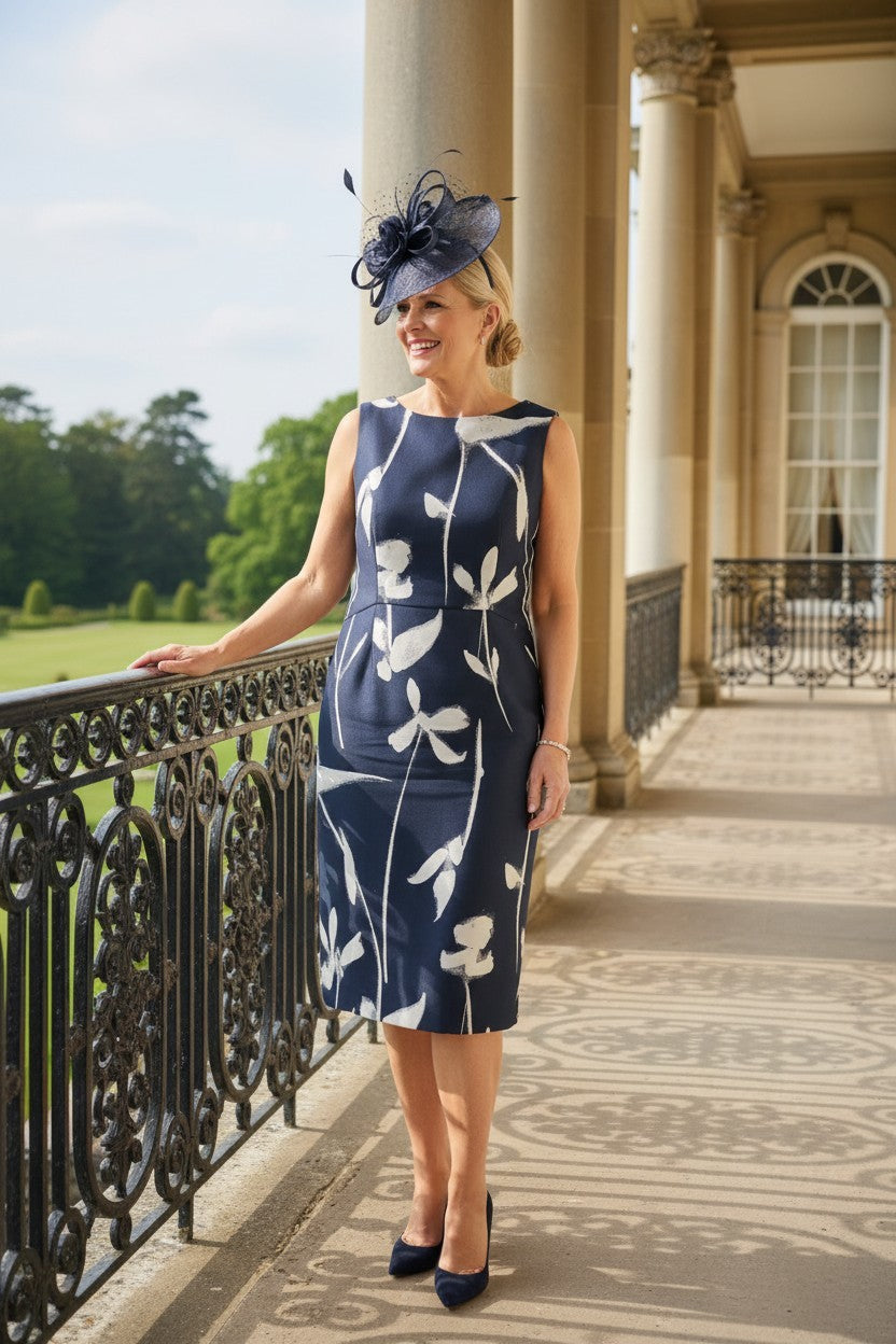 Navy mother of the bride dress by fely campo with matching navy fascinator, photographed in a formal garden setting.
