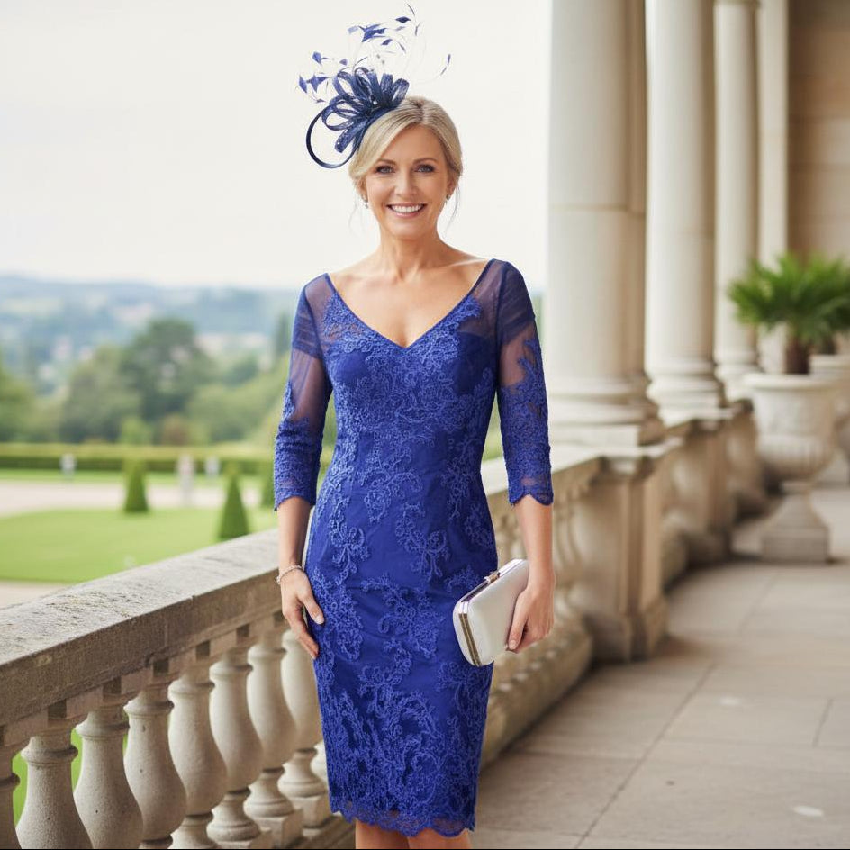 Woman in a irresistible cobalt blue mother of the bride dress with a decorative fascinator standing on a balcony with columns and greenery. Signature Dress