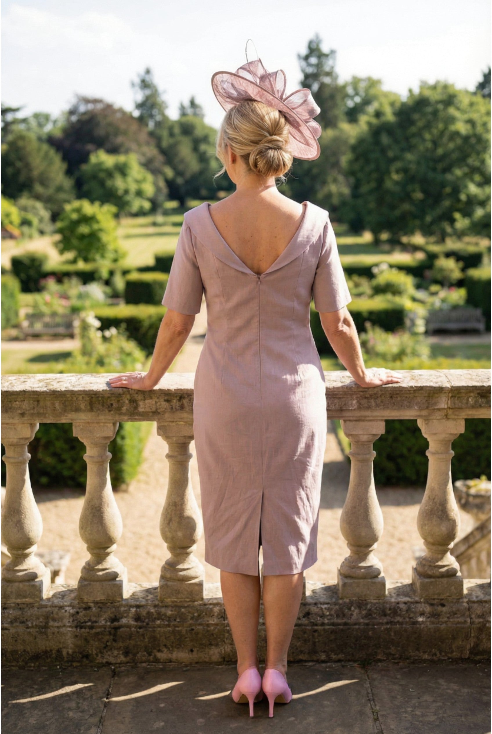 Woman in a ispirato light pink mother of the bride dress and pink fascinator  standing on a stone balcony with a garden view.
