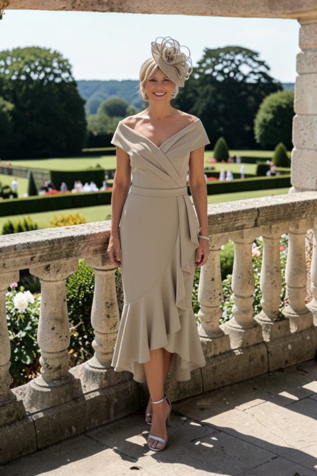 Taupe off‑the‑shoulder mother of the bride dress with fitted bodice and cascading hi‑lo frill hem, worn with matching fascinator on a stone terrace overlooking gardens.