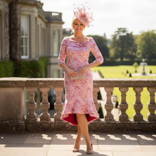 Woman in a pink Ispirato floral pink mother of the bride dress and pink fascinator hat standing in front of a classical building. Signature Dress.
