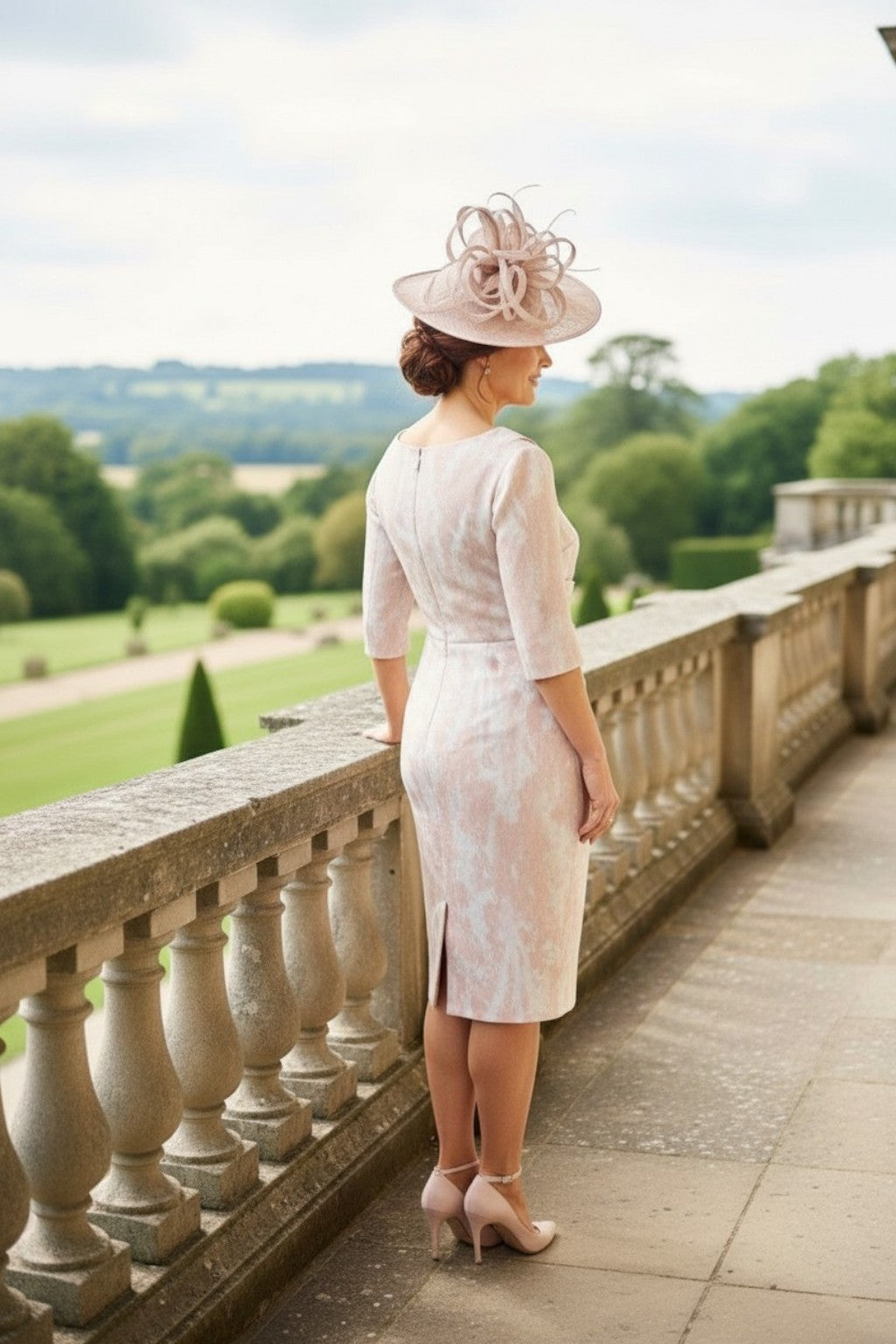 Blush pink mother of the bride dress with fitted silhouette, three‑quarter sleeves and matching wide‑brim hat, photographed from behind on a stone terrace overlooking gardens.