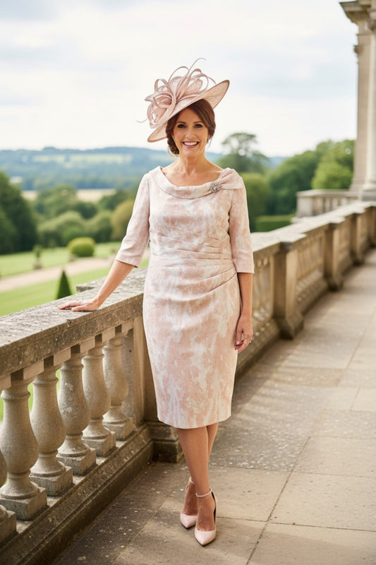 Blush pink mother of the bride dress with fitted silhouette, three‑quarter sleeves and matching wide‑brim hat, photographed from behind on a stone terrace overlooking gardens.