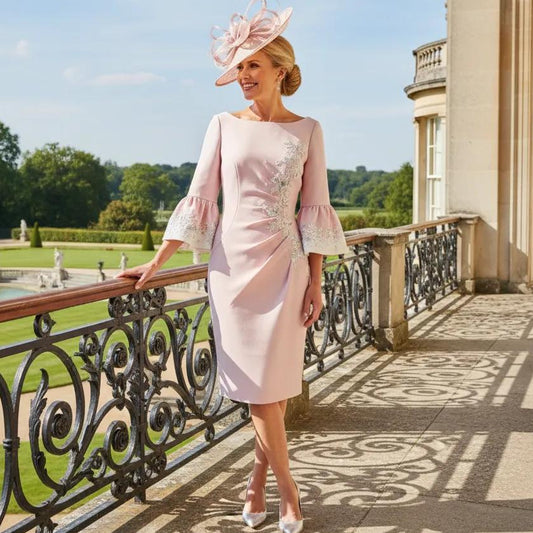 Woman in a light blush pink John Charles mother of the bride dress and matching pink fascinator hat standing on a balcony with a scenic background. Signature Dress.