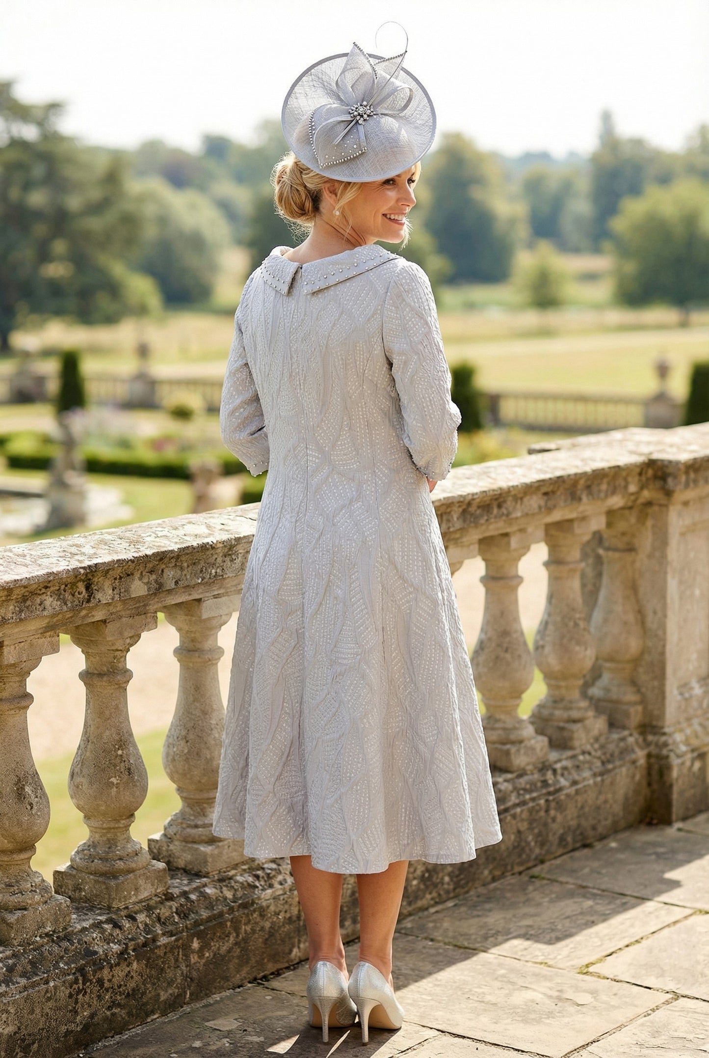 Back view of a woman in a A-Line light grey Lizabella mother of the bride dress with a large grey fascinator hat standing on a stone balcony.