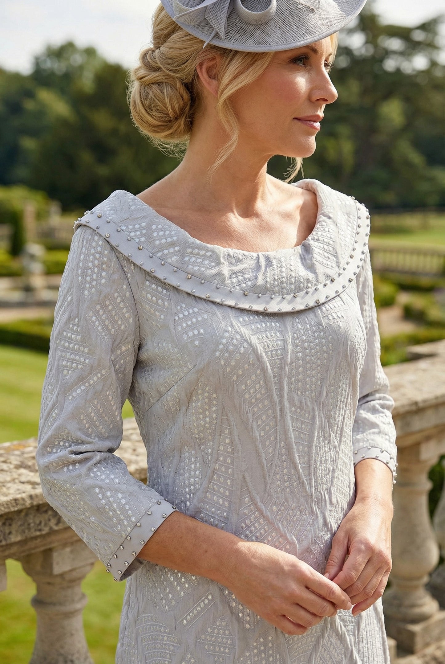 Close up image of a woman in a A-Line light grey Lizabella mother of the bride dress with a large grey fascinator hat standing on a stone balcony.