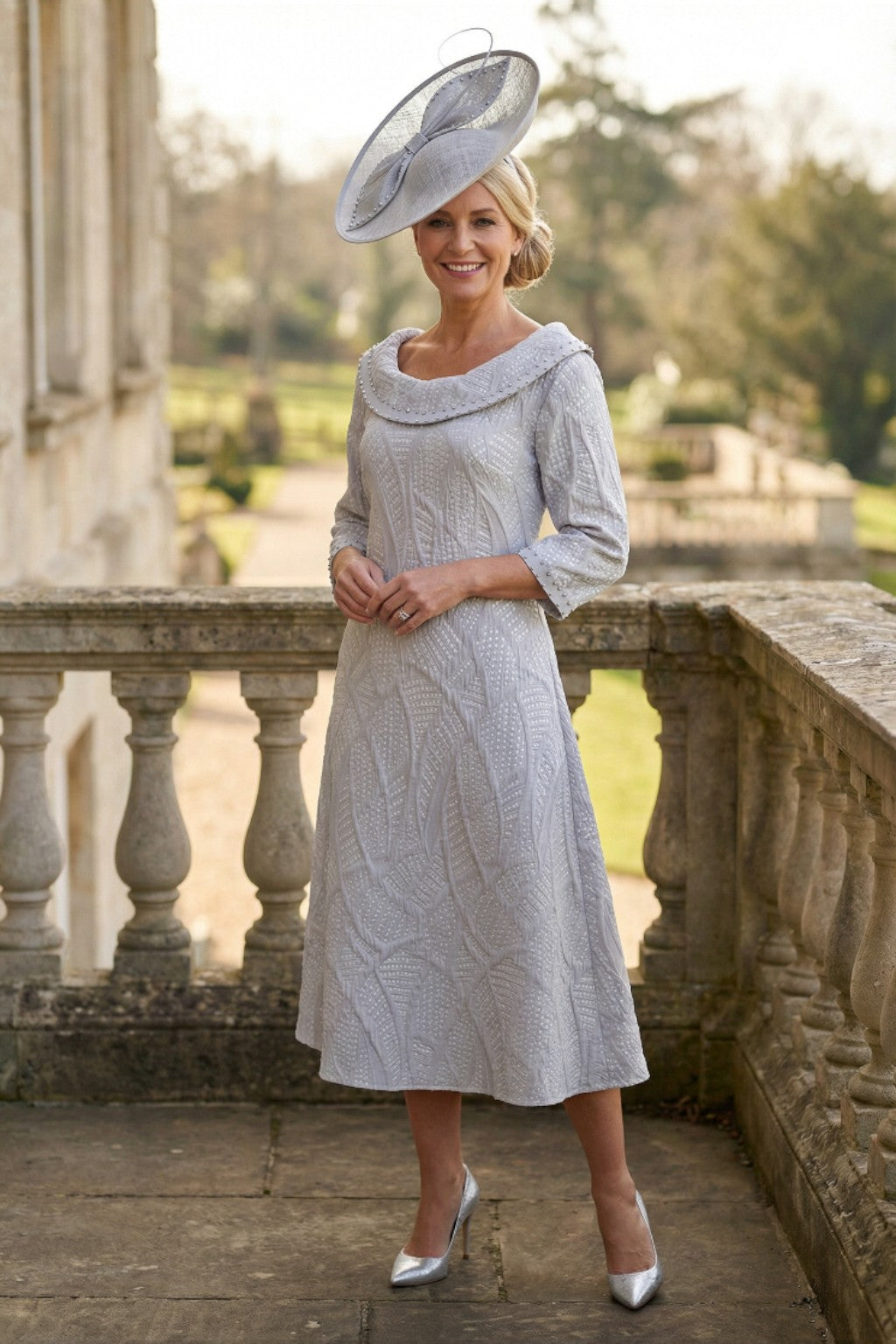 Woman in a A-Line light grey Lizabella mother of the bride dress with a large grey fascinator hat standing on a stone balcony.