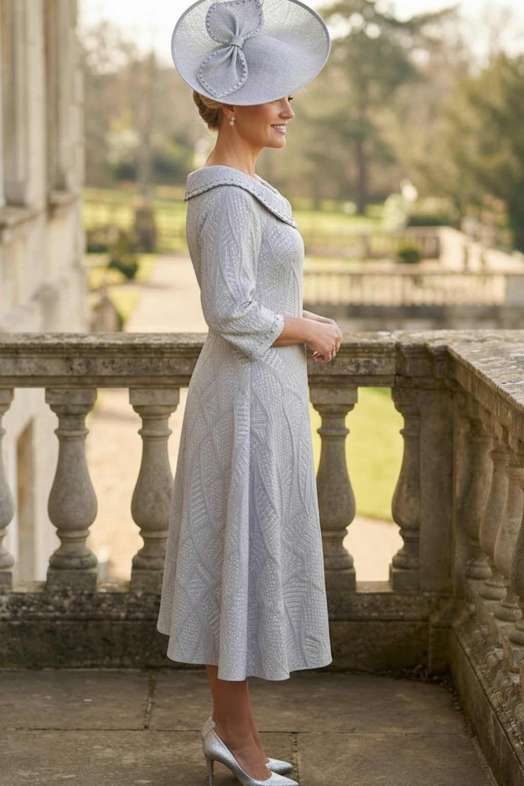 Side view of a woman in a A-Line light grey Lizabella mother of the bride dress with a large grey fascinator hat standing on a stone balcony.