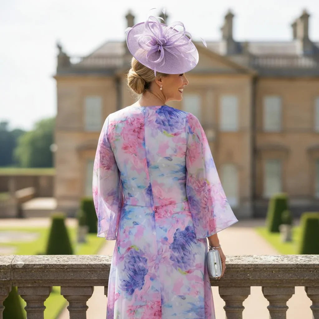 Woman in lizabella 2854 floral lilac mother of the bride dress and fascinator hat standing in a garden wedding setting showing the back of the dress.