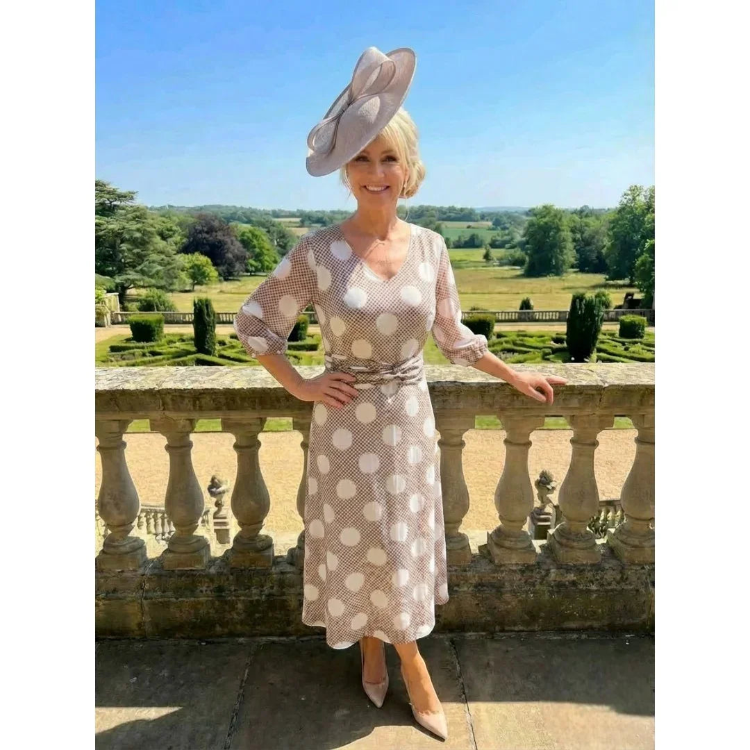 Woman in a Lizabella polka dot taupe mother of the bride dress and taupe fascinator hat standing on a stone balcony with a scenic background.