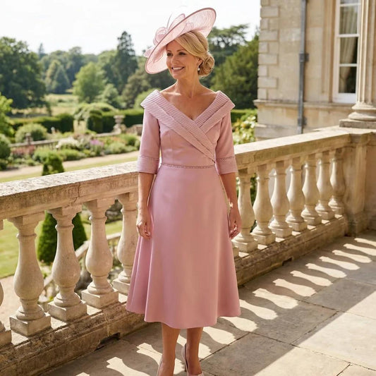 Light pink midi dress with pleated neckline and matching pink fascinator, worn on a balcony overlooking formal gardens.