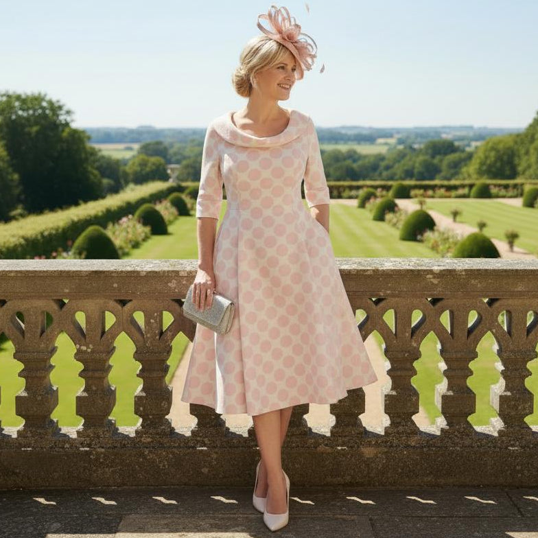 Woman in a Lizabella pink pokerdot spots mother of the bride dress and blush pink fascinator hat standing on a balcony with a scenic view of greenery. Signature Dress.