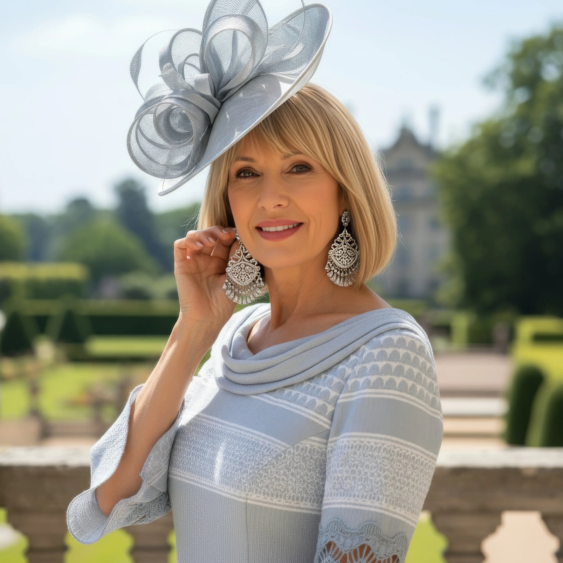 Woman in a striped silver mother of the bride dress and hat standing on a balcony with a garden and building in the background