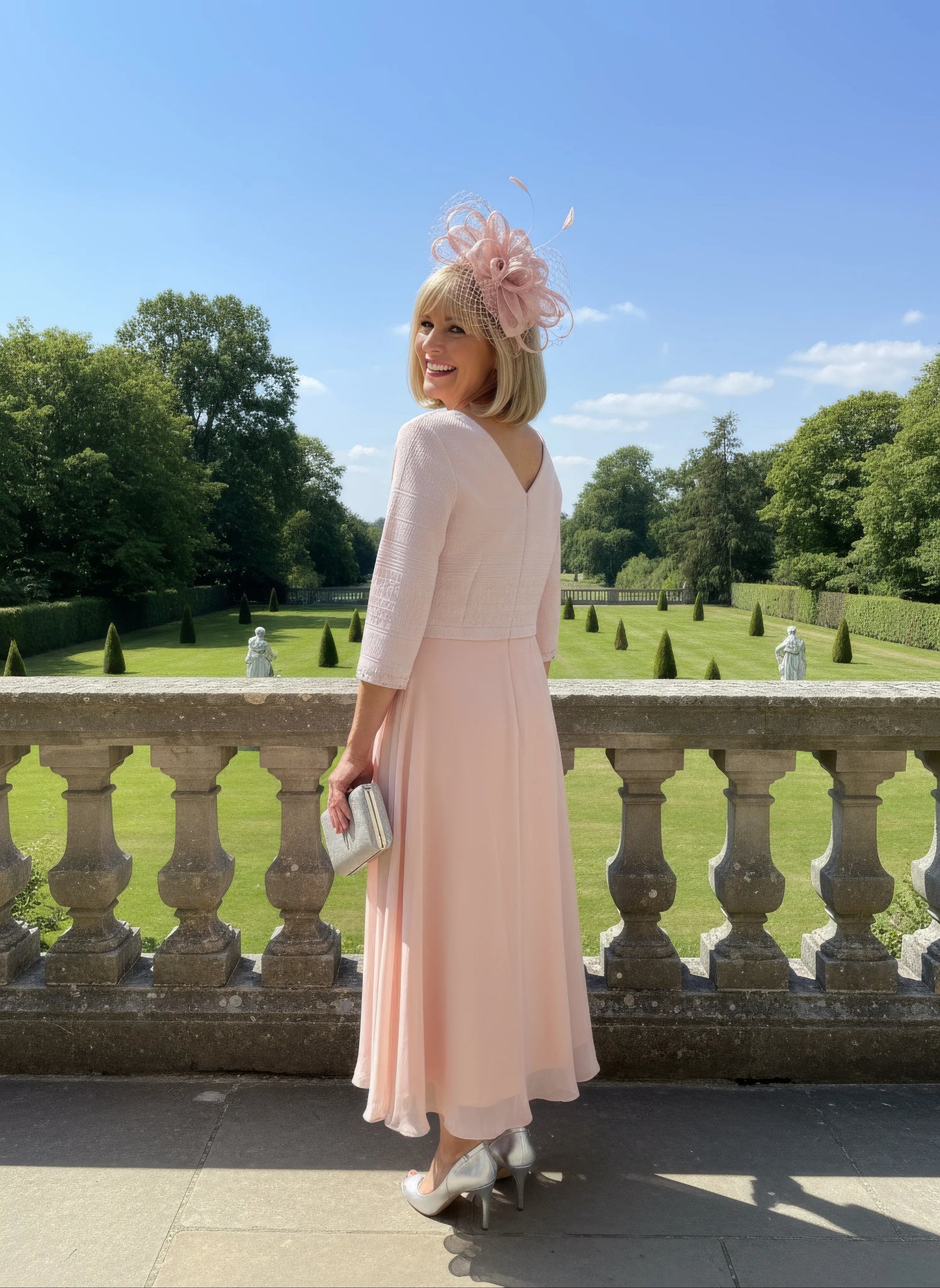 Woman in a light pink Mother of the bride dress by Lizabella with a decorative hat standing on a balcony with a scenic background.