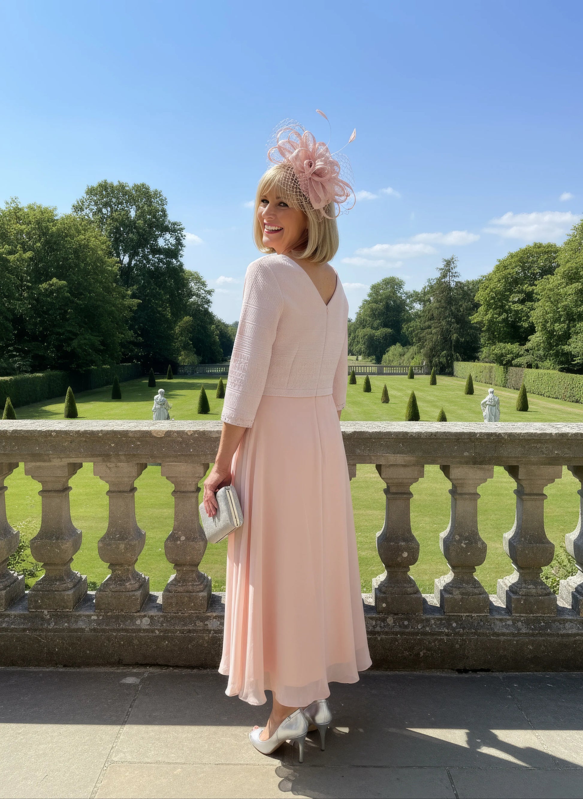 Woman in a light pink Mother of the bride dress by Lizabella with a decorative hat standing on a balcony with a scenic background.