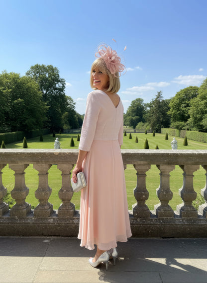 Woman in a light pink Mother of the bride dress by Lizabella with a decorative hat standing on a balcony with a scenic background.