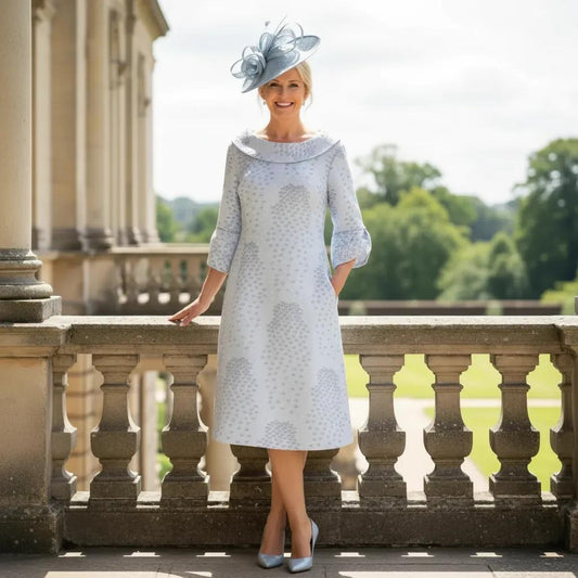 Woman in a Lizabella light blue A line mother of the bride dress and  pale blue fascinator hat standing on a balcony with greenery in the background. Signature Dress.
