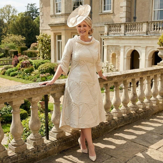 Woman in a Lizabella champagne mother of the bride dress and fascinator standing on a stone balcony. Signature Dress.