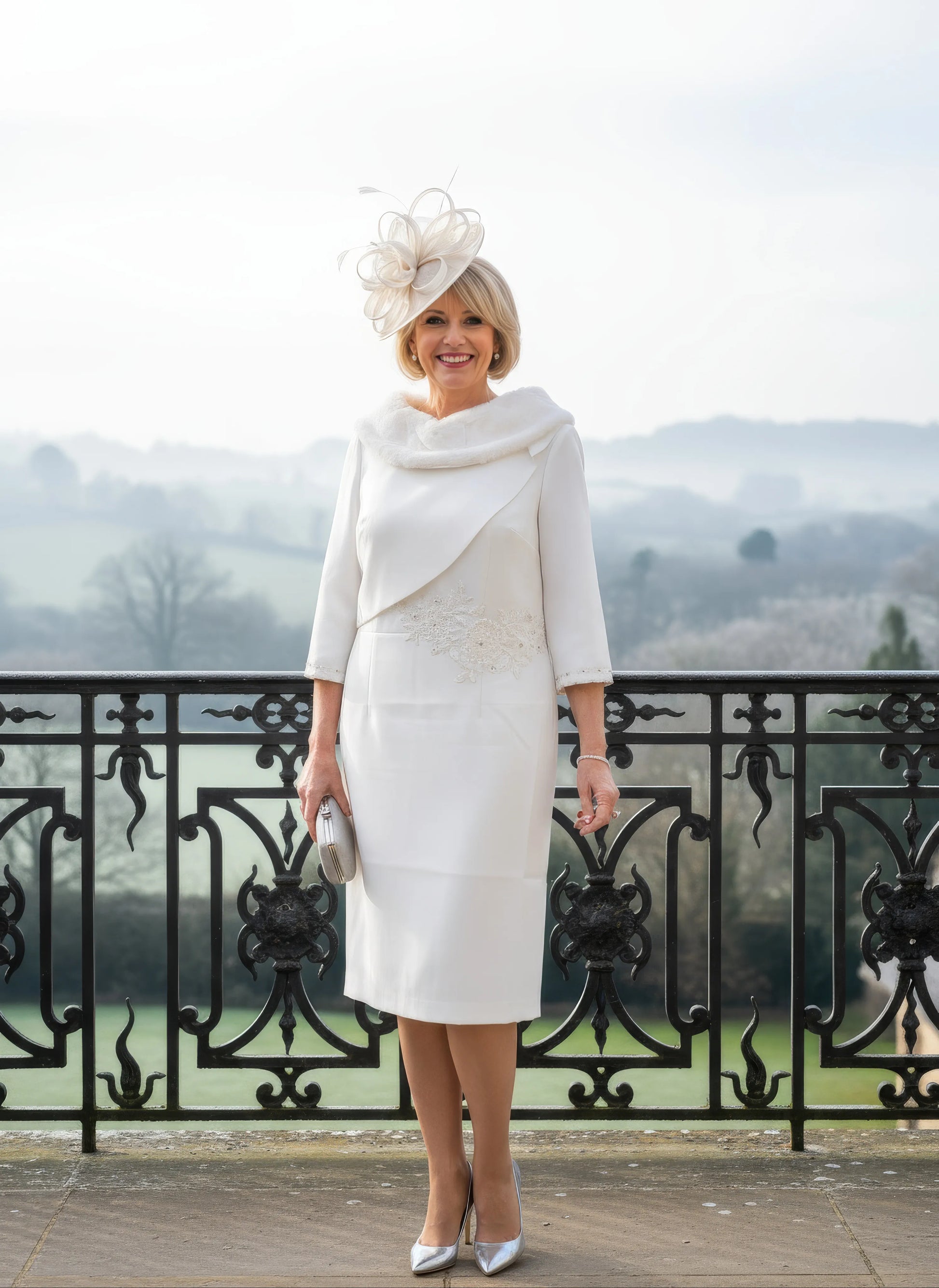 Woman in a white fur bridal dress with a matching hat standing on a balcony with a scenic background.
