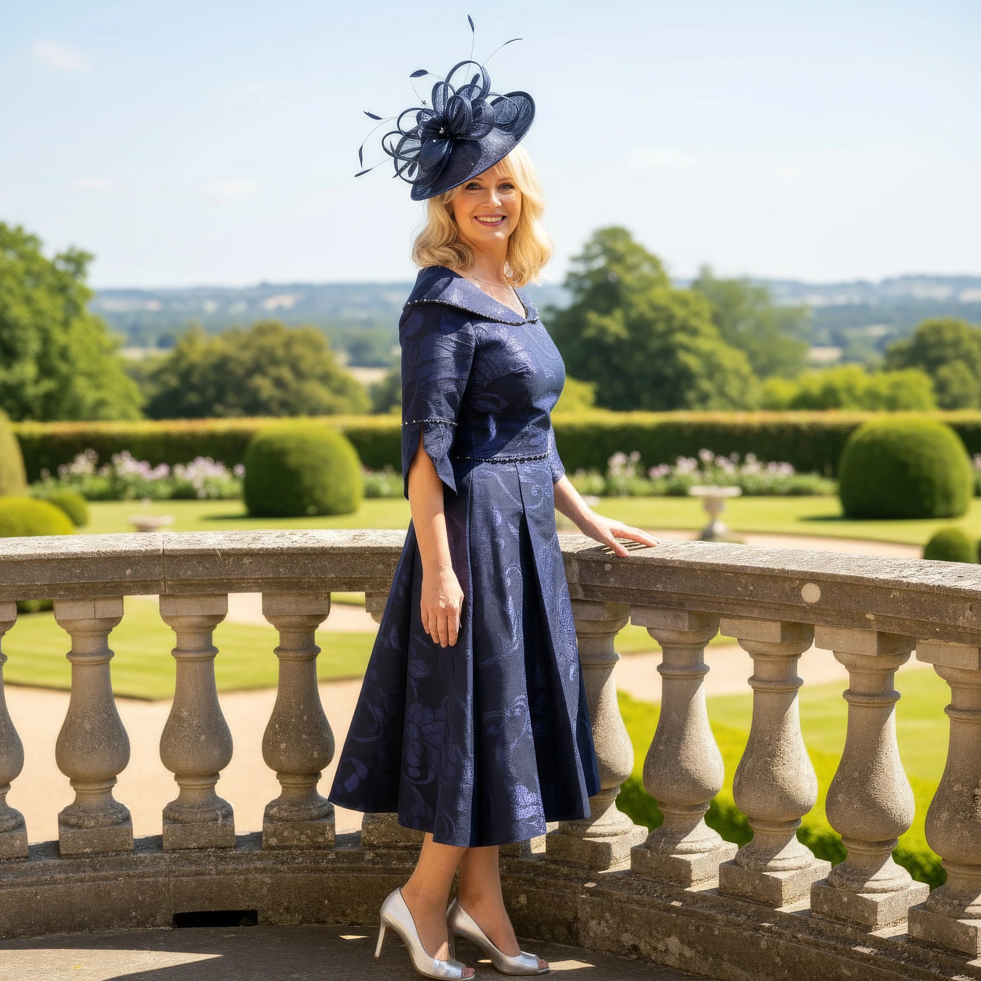 Woman in a navy blue Lizabella Mother of the bride dress with a matching hat standing on a balcony with a scenic background.