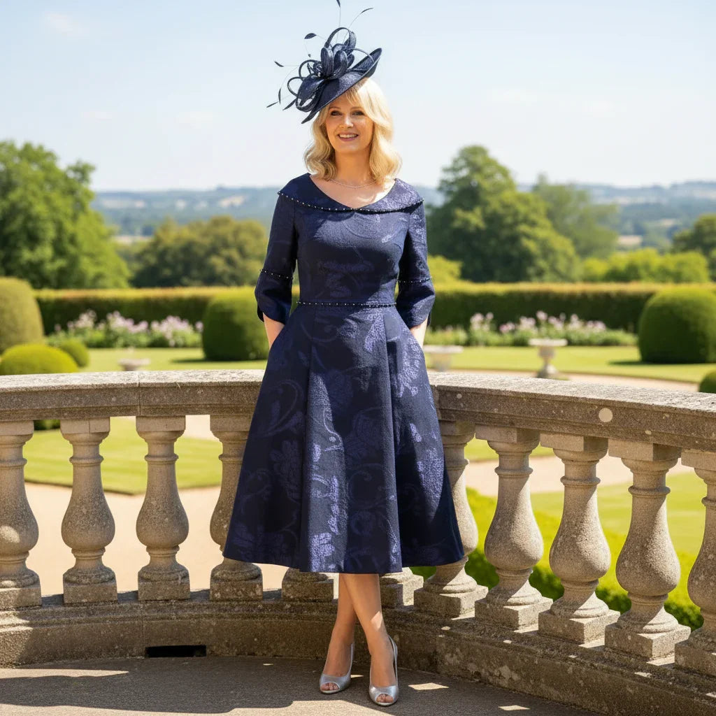 Woman in a navy blue Lizabella Mother of the bride dress with a matching hat standing on a balcony with a scenic background.