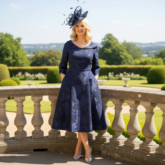 Woman in a navy blue Lizabella Mother of the bride dress with a matching hat standing on a balcony with a scenic background.