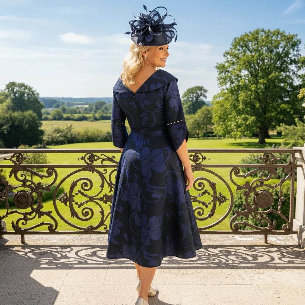Woman in a navy blue Lizabella Mother of the bride dress with a matching hat standing on a balcony with a scenic background.