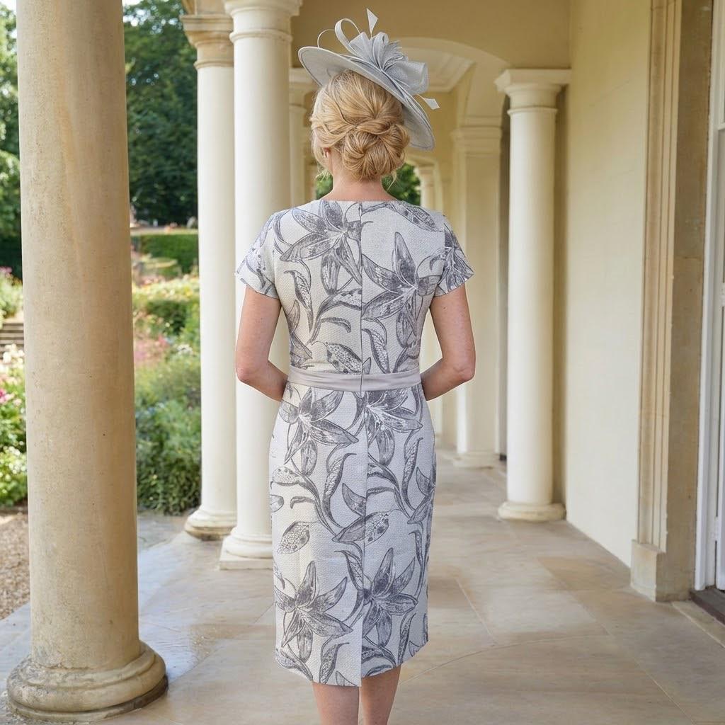 Woman in a floral Lizabella Silver 2 piece mother of the bride dress and matching jacket with a silver bespoke fascinator by Signature Dress standing in front of a large building with gardens. Signature Dress.