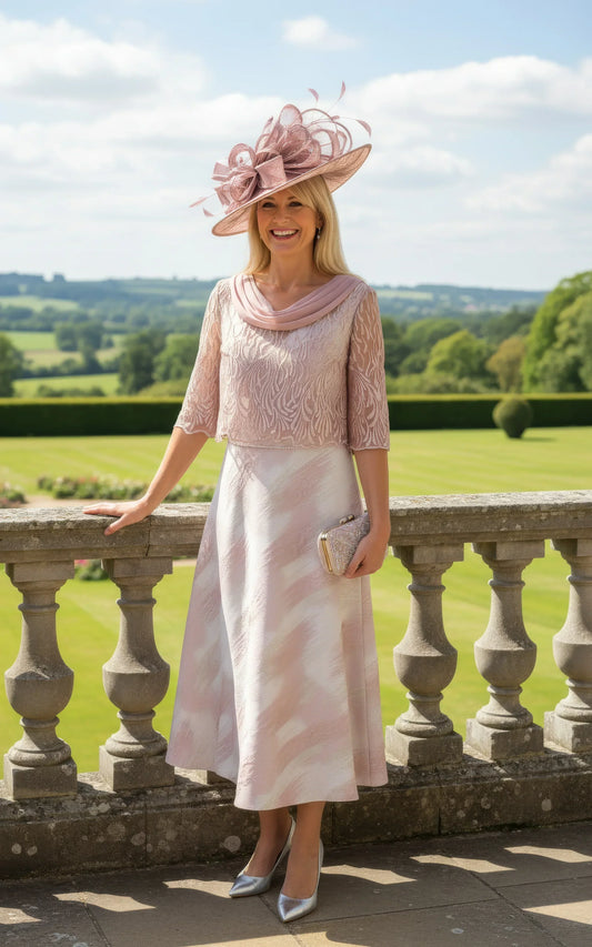 Woman in a pink Lizabella Mother of the bride dress and hat standing on a stone balcony with a scenic background