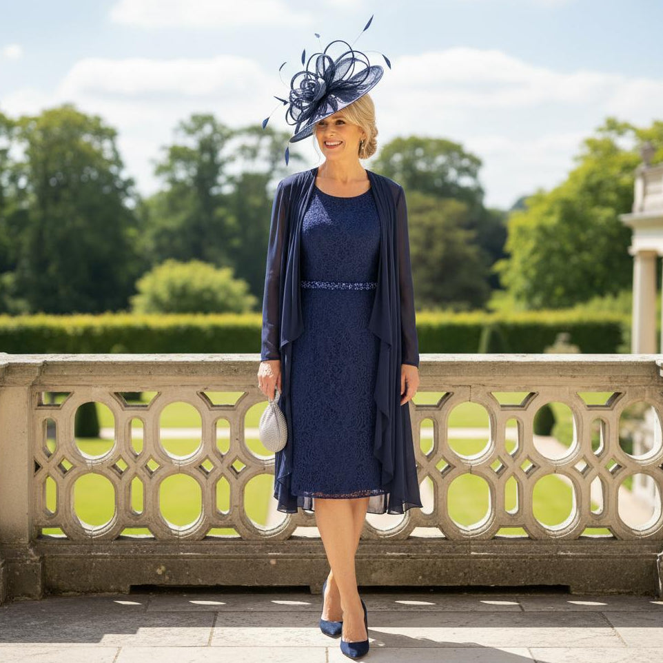 Woman in a mascara navy mother of the bride dress and matching jacket with a navy fascinator hat, standing outdoors. Signature Dress