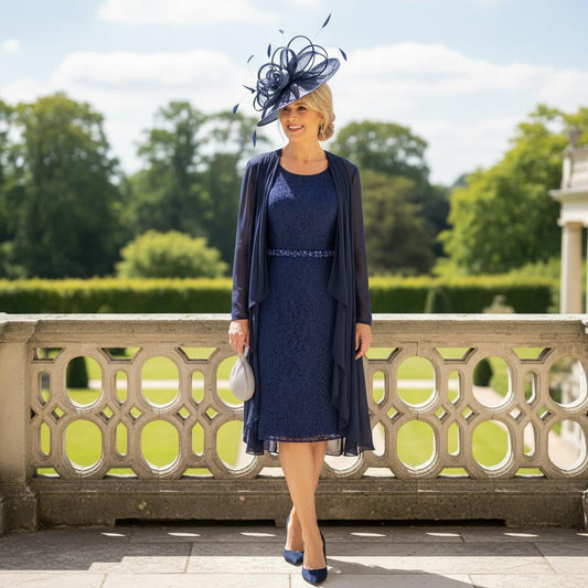 Woman in a mascara navy mother of the bride dress and matching jacket with a navy fascinator hat, standing outdoors. Signature Dress