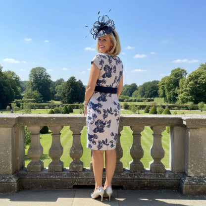 Woman in a floral dress with a hat standing on a stone balcony overlooking a garden.