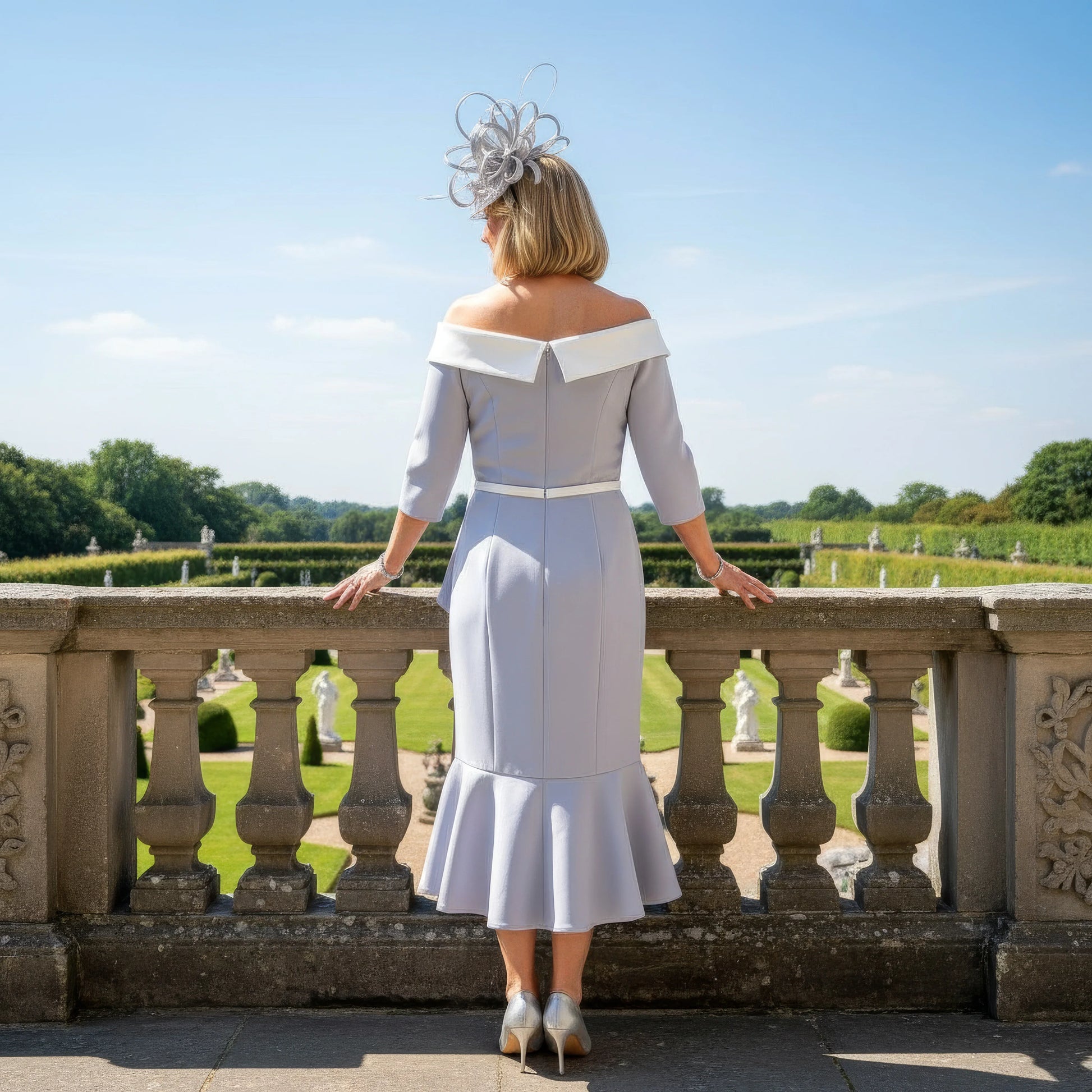 Woman in a formal Veni Infantino outfit with a large hat standing on a balcony with a scenic background