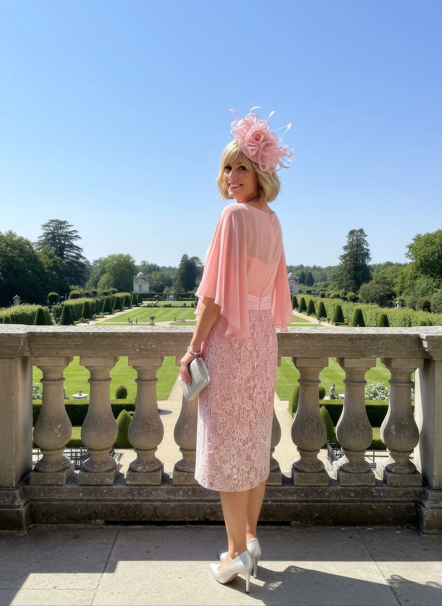 Woman in a ombre pink mother of the bride dress and pink fascinator standing on a stone balcony with a garden view.