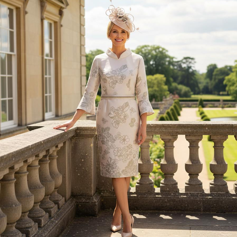 Woman in a formal Veni infantino gold mother of the bride dress and fascinator standing on a balcony with a scenic background. Signature Dress.